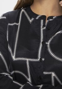 Laivastonsininen paita, jossa on geometrinen valkoinen kuvio, pehmeä tekstuuri, pyöristetty kaula-aukko ja mustat napit. Lähikuva kankaan yksityiskohdista.