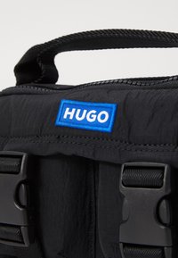 Black fabric bag with textured surface, featuring a blue and white "HUGO" logo. Includes black buckles and a sturdy handle.