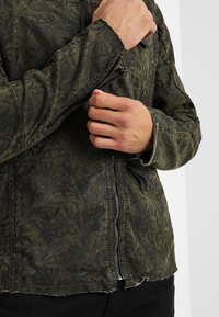 Kaki jacka med en texturerad bladmönster, med dragkedja fram och fransad fåll. Tyget verkar mjukt med en lätt sliten utseende.