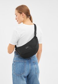 Borsa a tracolla nera realizzata in tessuto morbido, design curvo, tracolla regolabile e una patch con logo sul davanti, indossata sopra una maglietta bianca e jeans blu.