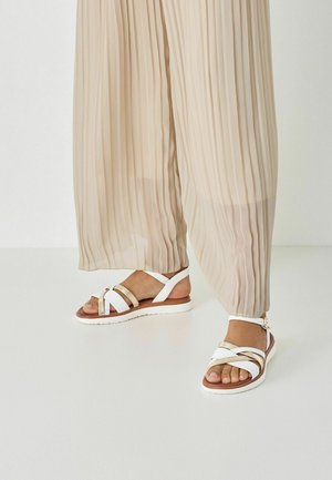 Bílé sandály s překříženými pásky a zlatým detailem na hnědé podrážce. Béžové plisované kalhoty částečně zakrývají boty. Hladká textura.