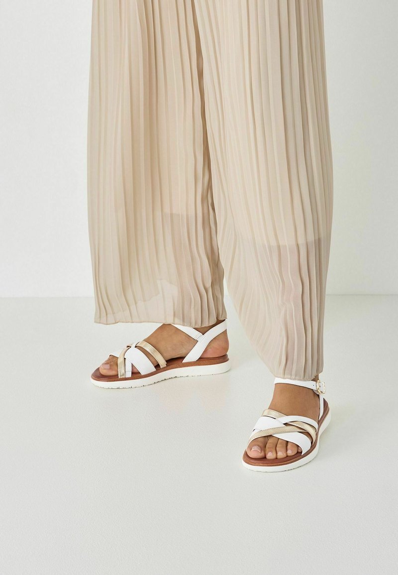 Sandales blanches à brides croisées avec un accent doré sur une semelle marron. Pantalon plissé beige couvrant partiellement les chaussures. Texture lisse.