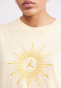 Person, die ein cremefarbenes Hemd mit gelbem Sonnenstrahl-Design und einer Silhouette eines Basketballspielers in der Mitte trägt, mit einem kleinen Herz-Tattoo am Hals.