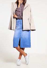 Beige ullblandad kappa, blå denimkjol med frontslits, och vita sneakers; mönstrad mörk skjorta under med volangdetaljer.