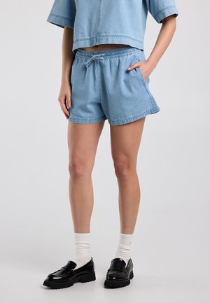 Personne portant un short en jean bleu clair, un top court assorti, des chaussettes blanches et des mocassins à plateforme noirs sur un fond uni.