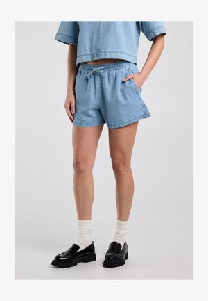 Personne portant un short en jean bleu clair, un top court assorti, des chaussettes blanches et des mocassins à plateforme noirs sur un fond uni.