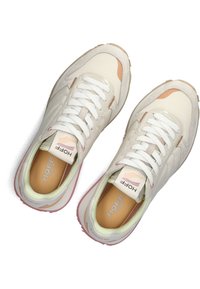 Zapatillas de deporte beige claro con cordones blancos, detalles en ante y un diseño curvado. Presentan un collar acolchado y una etiqueta de logo colorida en la lengüeta.