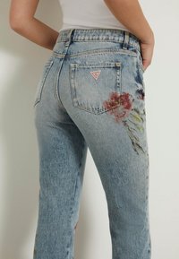 Jeans de mezclilla azul claro con bordado floral en rojo y verde en la pierna izquierda, que cuentan con un parche triangular del logo "Guess" en el bolsillo trasero.