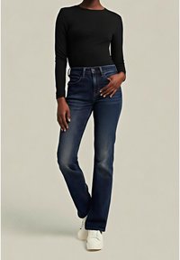 Personne portant un haut à manches longues noir, un jean droit bleu foncé et des baskets blanches, debout devant un fond neutre.