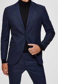 Mężczyzna poprawiający guzik na dopasowanej, ciemnoniebieskiej marynarce, noszonej na czarnym golfie, pokazany od szyi do górnej części ud na jasnym tle.