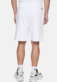 Shorts en coton blanc avec une taille élastique, des poches latérales et un petit patch en logo, associés à des baskets blanches et des chaussettes.
