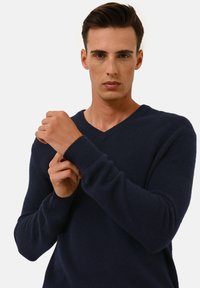 Navyblauer V-Ausschnitt-Pullover aus weichem, strukturiertem Material; mit gerippten Bündchen und entspannter Passform. Das Model passt die Ärmel an.