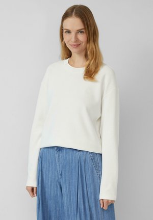 Vrouw met lichtbruin haar die een witte lange mouwen trui en blauwe geplooide broek draagt tegen een effen lichtgrijze achtergrond.