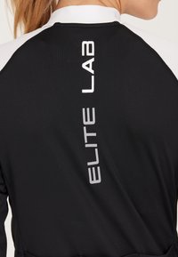 Schwarzes Sporttop aus strukturiertem Stoff mit weißen Akzenten und vertikaler "ELITE LAB"-Markierung entlang des Rückens.