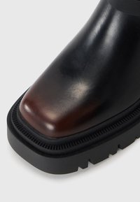 Sort læder ankelstøvle med en rund tå og brun poleret detalje fortil. Har en kraftig sort gummisål med tekstureret detaljering.