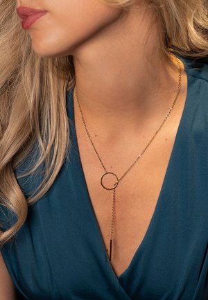 Gouden hangerketting met een cirkelring en een verticale staaf aan een ketting, gedragen over een blauwe kledingstuk met een v-hals ontwerp.