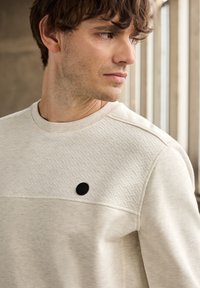 Světlý béžový svetr s texturovaným horním panelem, žebrovaným límcem a černým tlačítkem s logem. Materiál vypadá měkký s jemným vzorem.