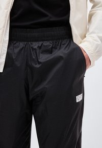 Μαύρο καρό παντελόνι με ελαστική μέση, cropped λευκή μανσέτα σακακιού και χέρι στην τσέπη του παντελονιού που φαίνεται σε ένα άτομο.