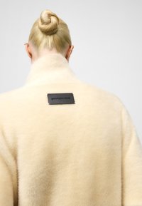 Crèmekleurige wollen jas met een zachte textuur, hoge kraag en een zwart merkembleem op de achterkant. Minimalistisch ontwerp, geen zichtbare hardware.