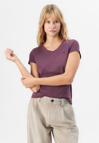 Blonde Frau in einem lila kurzärmeligen V-Ausschnitt-Shirt und beigen hochgezogenen Hosen, die mit verschränkten Armen vor einem weißen Hintergrund posiert.