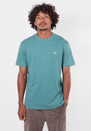 Homme aux cheveux bouclés portant un t-shirt bleu sarcelle à manches courtes et un pantalon marron, se tenant devant un fond blanc uni.