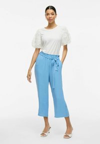 Witte top met gestructureerde pofmouwen, gecombineerd met blauwe cropped broeken met een strikband en witte open teen hakken.