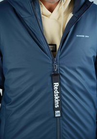 Doudoune bleu marine avec zip intégral, capuche à cordon de serrage et étiquette logo « Redskins 1984 ». Texture lisse avec peu de détails.