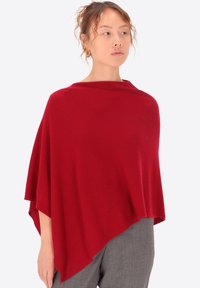 Poncho rosso lavorato a maglia con una silhouette rilassata e drappeggiata. Presenta una texture morbida, scollo ampio e orlo asimmetrico. Indossato sopra pantaloni grigi.