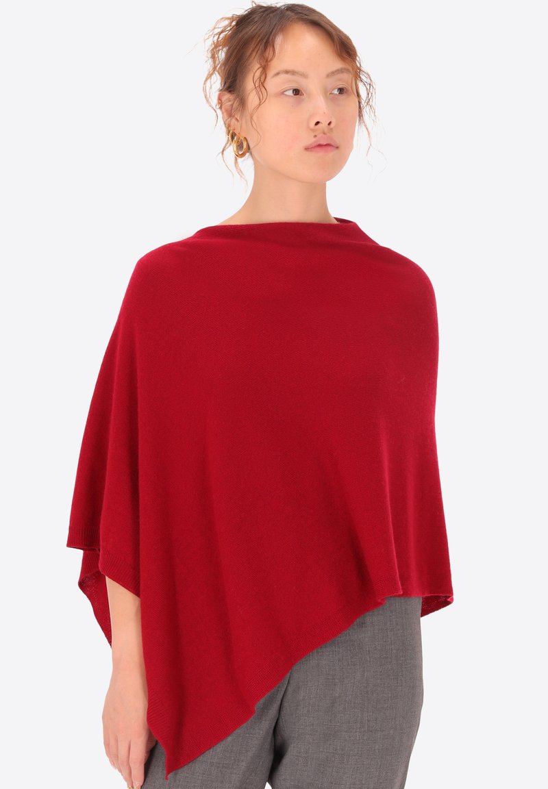 Poncho rosso lavorato a maglia con una silhouette rilassata e drappeggiata. Presenta una texture morbida, scollo ampio e orlo asimmetrico. Indossato sopra pantaloni grigi.