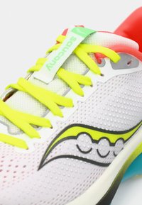 Zapatilla de running con una parte superior de malla en blanco y rosa, cordones amarillos y acentos grises. Presenta un talón colorido y un detalle del logo.