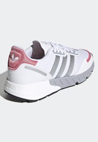 Vit träningsskor med grå ränder, texturerad ovandel, rosa detaljer vid hälen och en tjock vit mellansula med svart sulmönster.