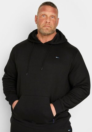 Homme aux cheveux courts et à la barbe, portant un sweat à capuche noir avec les mains dans la poche avant, regardant légèrement vers le haut sur un fond uni.