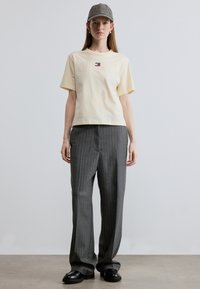 Ανοιχτό κίτρινο βαμβακερό μπλουζάκι με κοντά μανίκια και μικρό λογότυπο στο στήθος, συνδυασμένο με γκρι παντελόνι καμπάνα με λεπτές ραβδώσεις και μαύρα παπούτσια.