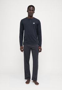 Camisa oscura de manga larga con placket de botones y un pequeño logo, combinada con pantalones de pijama oscuros con un diseño geométrico.
