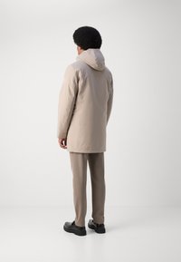 Parka beige con capucha y un acabado suave y mate, que presenta un corte recto y una parte trasera más larga. Combinada con pantalones beige y zapatos negros.