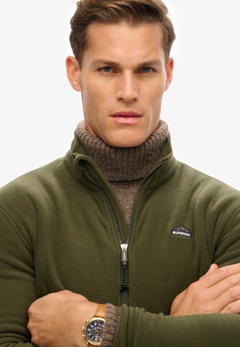 Veste en polaire verte avec zip et logo, superposée à un col roulé côtelé marron. Montre dorée visible au poignet, bras croisés.