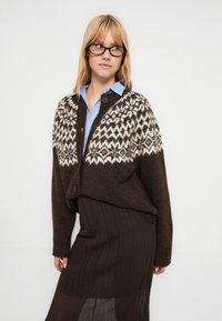Brown knitted cardigan with a patterned light top half, featuring three buttons and a loose fit. Worn over a ribbed brown skirt.