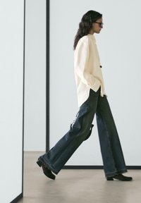 Camicia bianca a maniche lunghe con dettagli a bottoni abbinata a jeans a gamba larga blu scuro e stivaletti neri, su uno sfondo neutro.