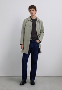 Jeune homme portant un trench coat gris clair sur un pull noir, un jean bleu foncé, une ceinture noire et des mocassins noirs, se tenant contre un mur uni.