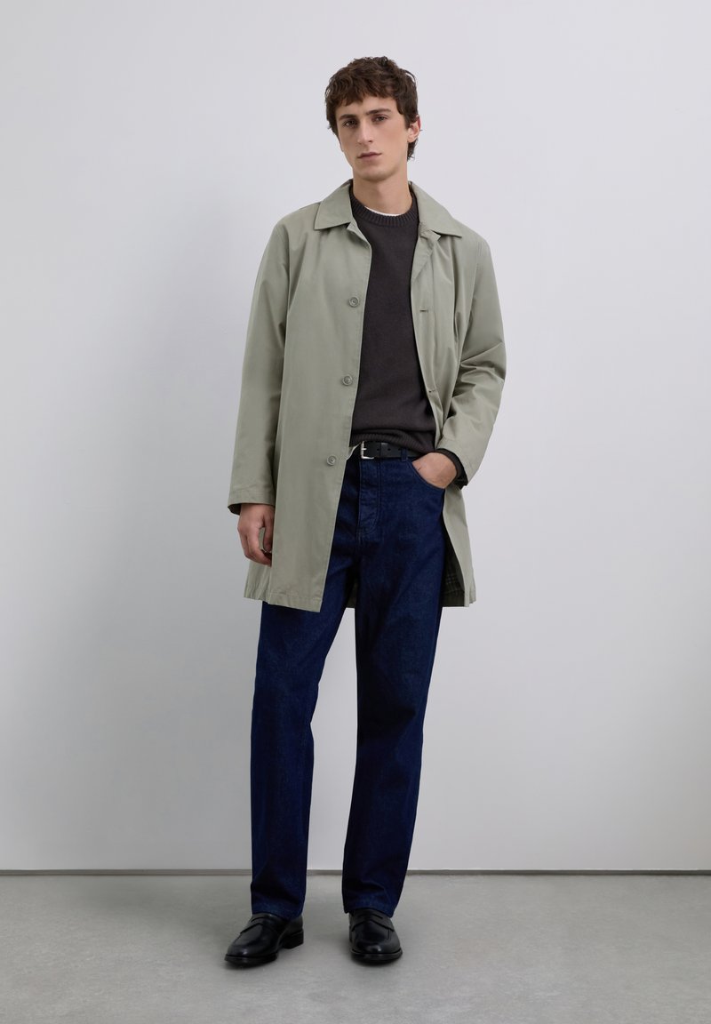 Jeune homme portant un trench coat gris clair sur un pull noir, un jean bleu foncé, une ceinture noire et des mocassins noirs, se tenant contre un mur uni.