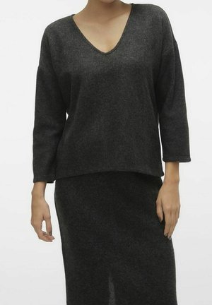 Femme portant un pull en maille côtelée gris foncé à col en V et une jupe assortie avec une fente à l’avant, debout sur un fond blanc uni.