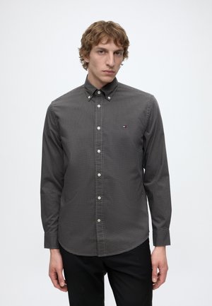 Jeune homme aux cheveux bouclés portant une chemise à carreaux noire et blanche boutonnée et un pantalon noir sur un fond blanc uni.
