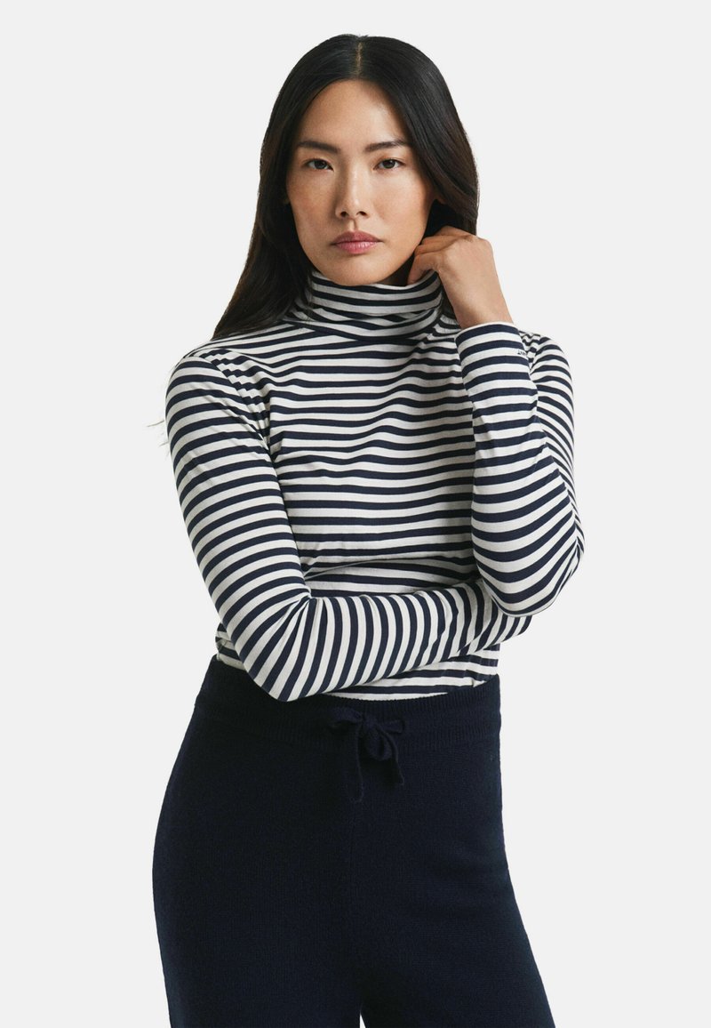 Vrouw met lang donker haar, gekleed in een zwart-wit gestreepte coltrui en zwarte broek, staand met een hand bij haar nek.