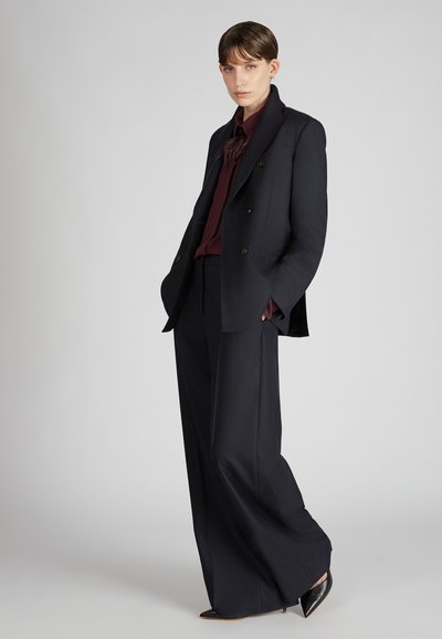 Persoon in wijd-uitlopende zwarte broek, dubbelzijdig zwarte blazer, bordeauxblauwe blouse en zwarte puntige hakken staat tegen een grijze achtergrond.