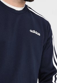 Sudadera azul marino de algodón con un diseño de tres rayas blancas a lo largo de los hombros y el logo blanco de Adidas en el pecho.
