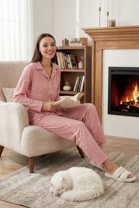 Frau in pinkem Schlafanzug liest ein Buch in einem beigen Sessel neben einem Kamin, neben ihr schläft eine weiße Katze auf einem Teppich.