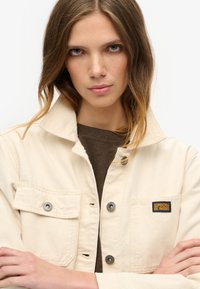 Beige canvasjakke med krave, trykknapper og to frontlommer. Har et blåt og gult "Superdry" logo-patch på brystet.