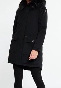 Sort isoleret parka-jakke med faux fur krave, frontlukning med knapper og lynlås, to store lommer, tekstureret stof og justerbare manchetter.