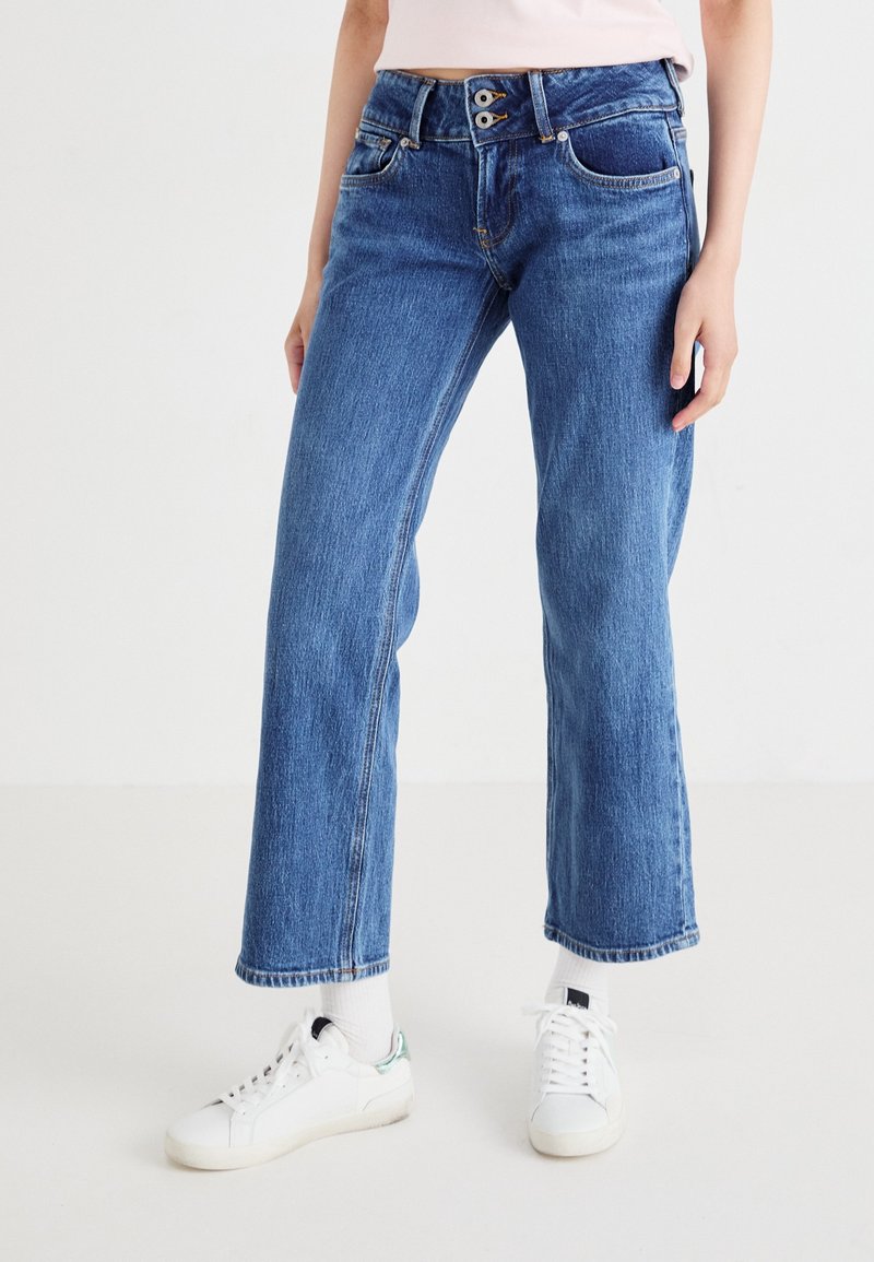 Jean en denim bleu à jambe large et taille haute, avec fermeture à deux boutons, poches avant et une texture légèrement décolorée. Porté avec des baskets blanches.