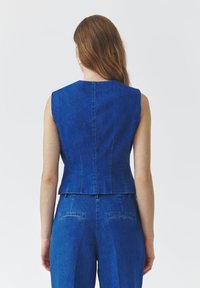 Modra jeans srajca brez rokavov z oprijetim dizajnom, visokim ovratnikom in čistimi šivi. Tkanina ima gladko teksturo in rahlo izbledi videz.